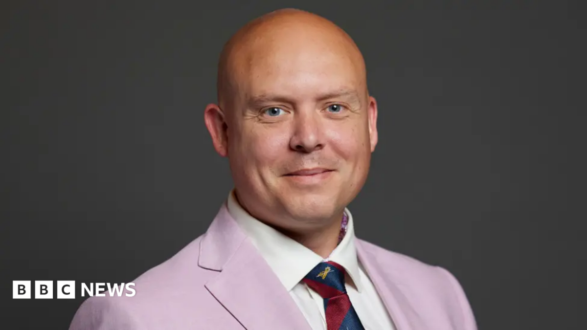 Head and shoulders shot of Cameron Thomas MP looking at the camera and smiling - wearing a pink suit, white shirt and red and blue stripped tie. 