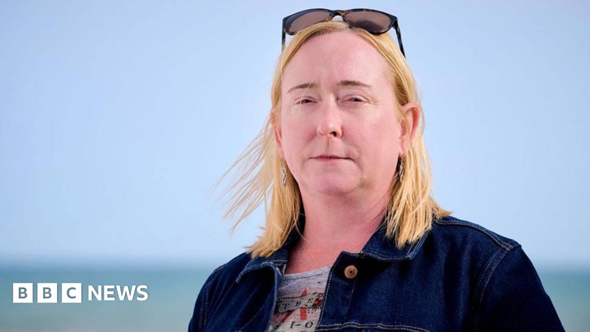 A woman with blond shoulder-length hair, standing on a pebble beach with the sea in the background. She is wearing a blue denim jacket. The sky is blue and cloudless.