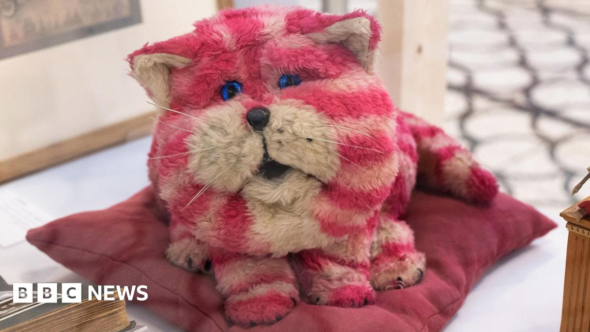 Bagpuss, a pink and white furry toy cat, sitting on a purple cushion