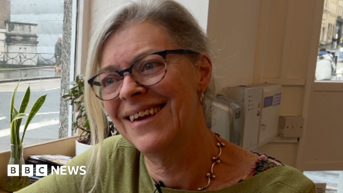 A headshot of Phoebe looking to the right of camera and smiling. There is a window to her right and she is wearing a green jumper and a chunky gold necklace and glasses.