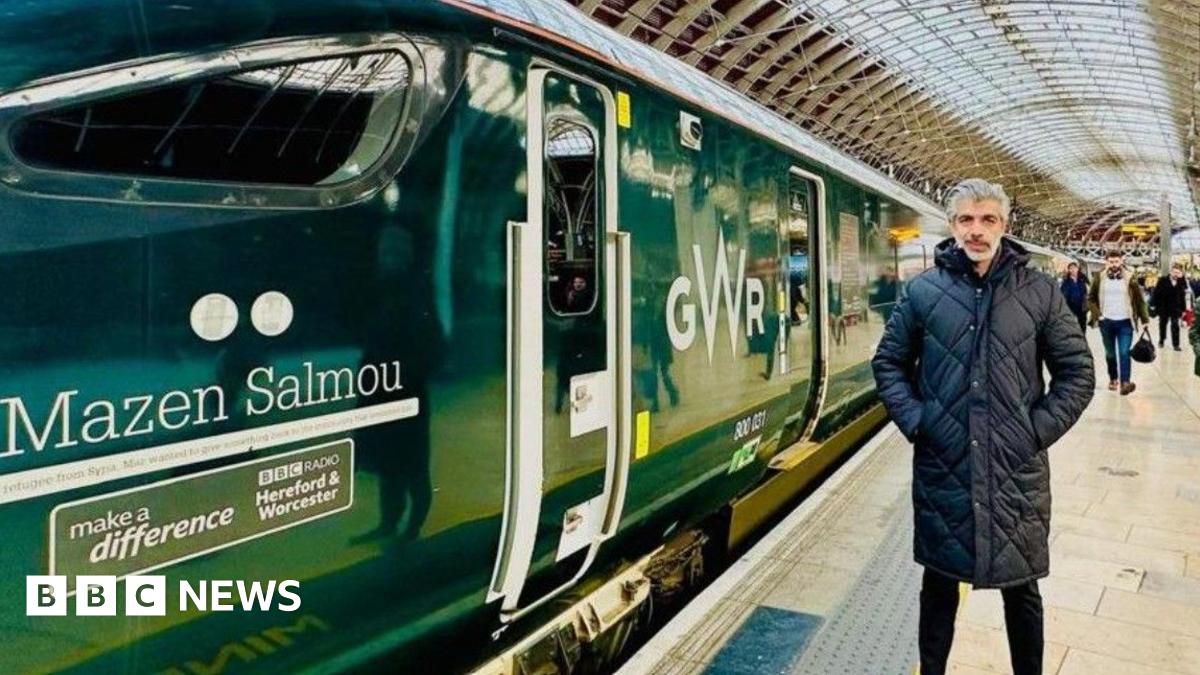 A man with grey hair is wearing a coat on the right hand side of the photo. The image is dominated by a green train at a railway station. On the left of the image the train has the words Mazen Salmou on it.