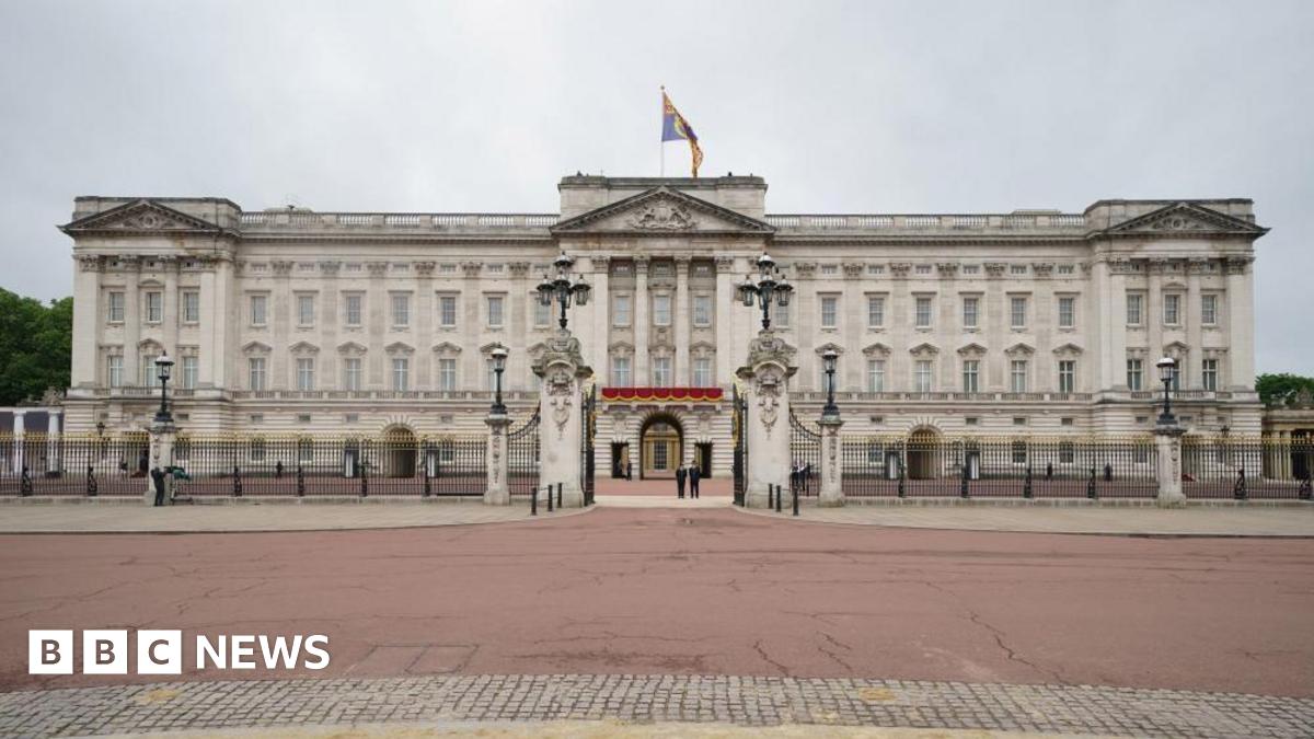 A frontal view of Buckingham Palace. The only people in the shot are two police officers standing just inside the palace's open gates. 