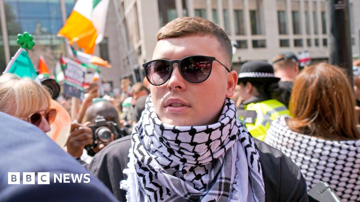 A close-up of a man in the midst of a crowd. He is wearing sunglasses and a black and white keffiyeh headdress around his neck like a scarf. A crowd is around him. You can see Irish tricolours in the background