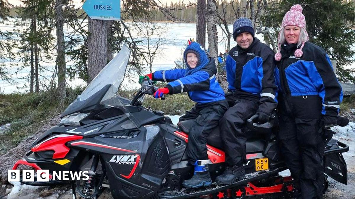 Wayne O'Neil and his son Finn sitting on a snowmobile with Lisa O'Neil standing next to them. They are all wearing snow suits and woolly hats, but there is no snow on the ground around them. There is a lake in the background.