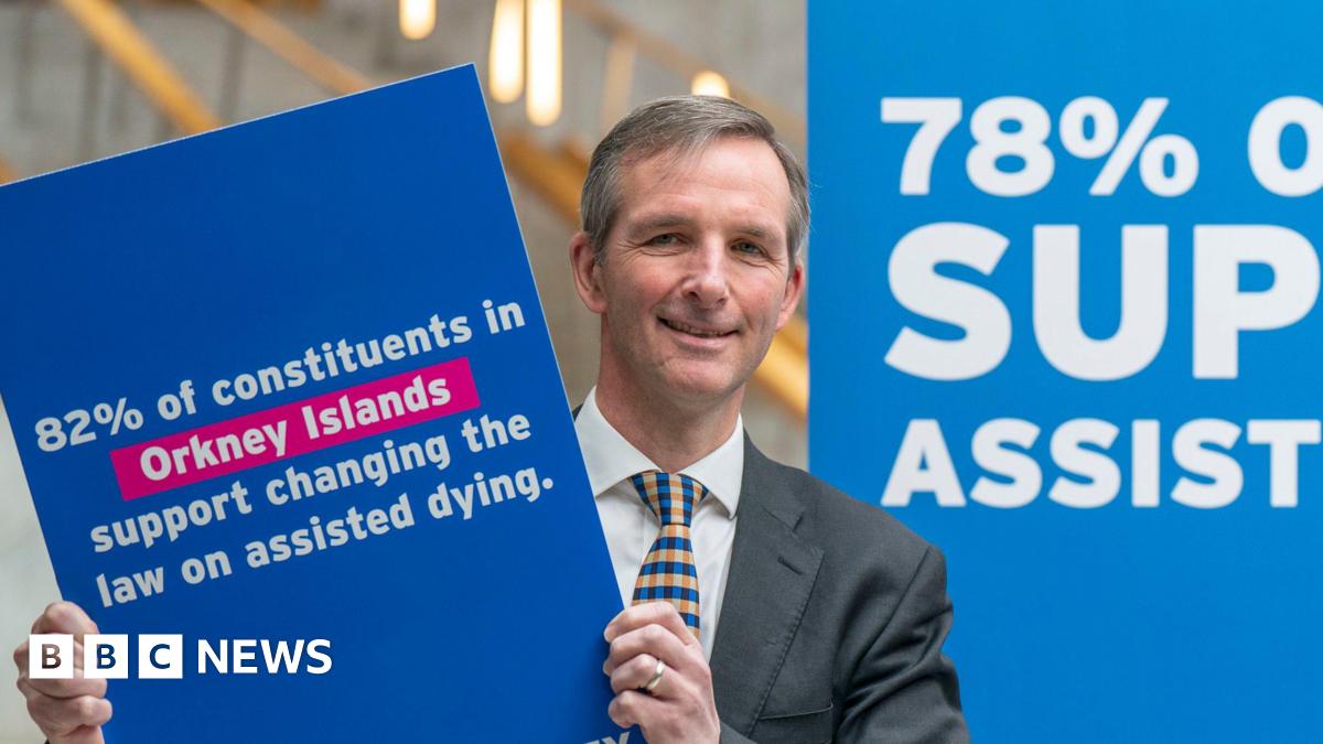 Liam McArthur - a man with brown hair and wearing a suit with a yellow and blue tie, holds up a placard saying people in the Orkney Islands support changing the law on assisted dying.