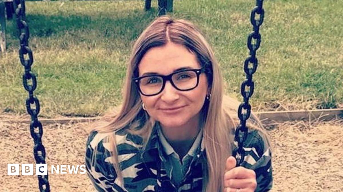 Isobella Knight with long blonde hair wearing large dark brown glasses. She is wearing a black and gold top and is holding onto the chain of a swing in a park. There is a dried grass landing area behind her and green grass beyond that.