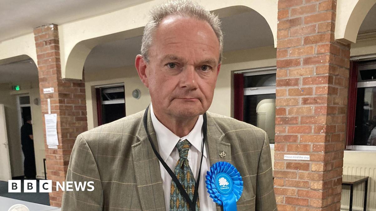 A man with short grey hair, a grey jacket, green tie and a blue Conservative Party rosette, stands in a hall with brick pillars and a white wall with windows behind him.