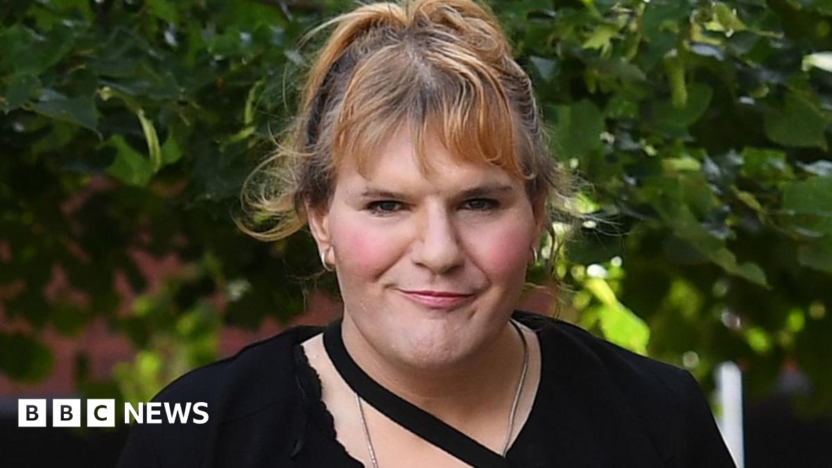 Katie Wallis facing the camera as she walks along a street. She is wearing a black dress with a black strap across her neck. She has her hair up.