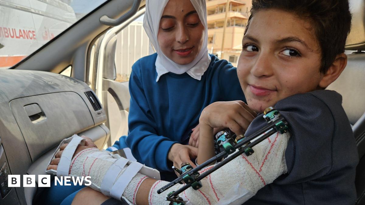 Adam al-Najjar sits in an ambulance with a brace around his arm wearing a dark t-shirt while his cousin, wearing blue and a white headscarf, tends to him.