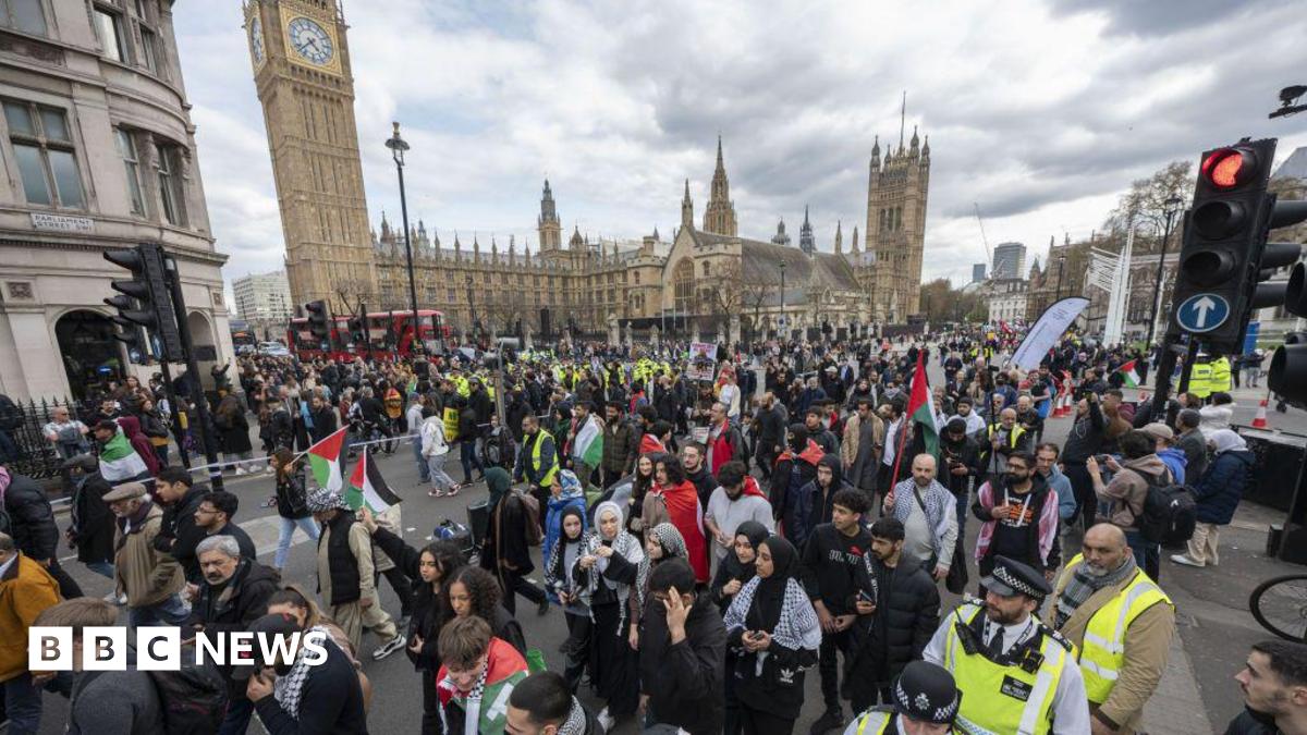 Large march in central London with the Houses of Parliament behind a crowd of marching protesters. Some are holding Palestinian flags