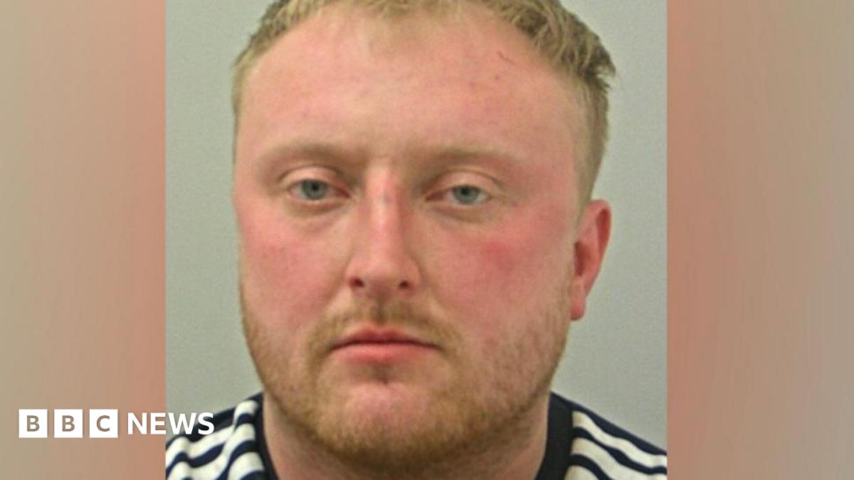 Zak Honeyman's police mugshot, showing a white man with rosy cheeks and pale eyes, short blond hair and a short beard. He is wearing a black and white striped top.