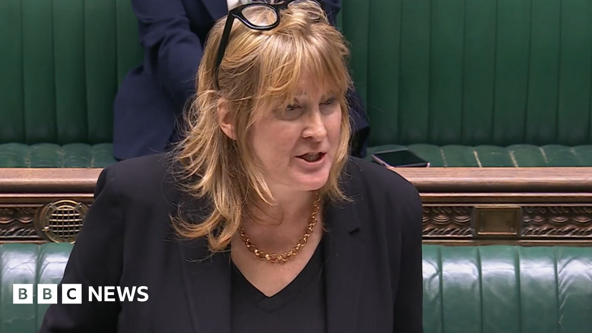 A woman with long fair hair stands at the Commons despatch box with green benches behind her. She is wearing a black suit and top, and a gold necklace.