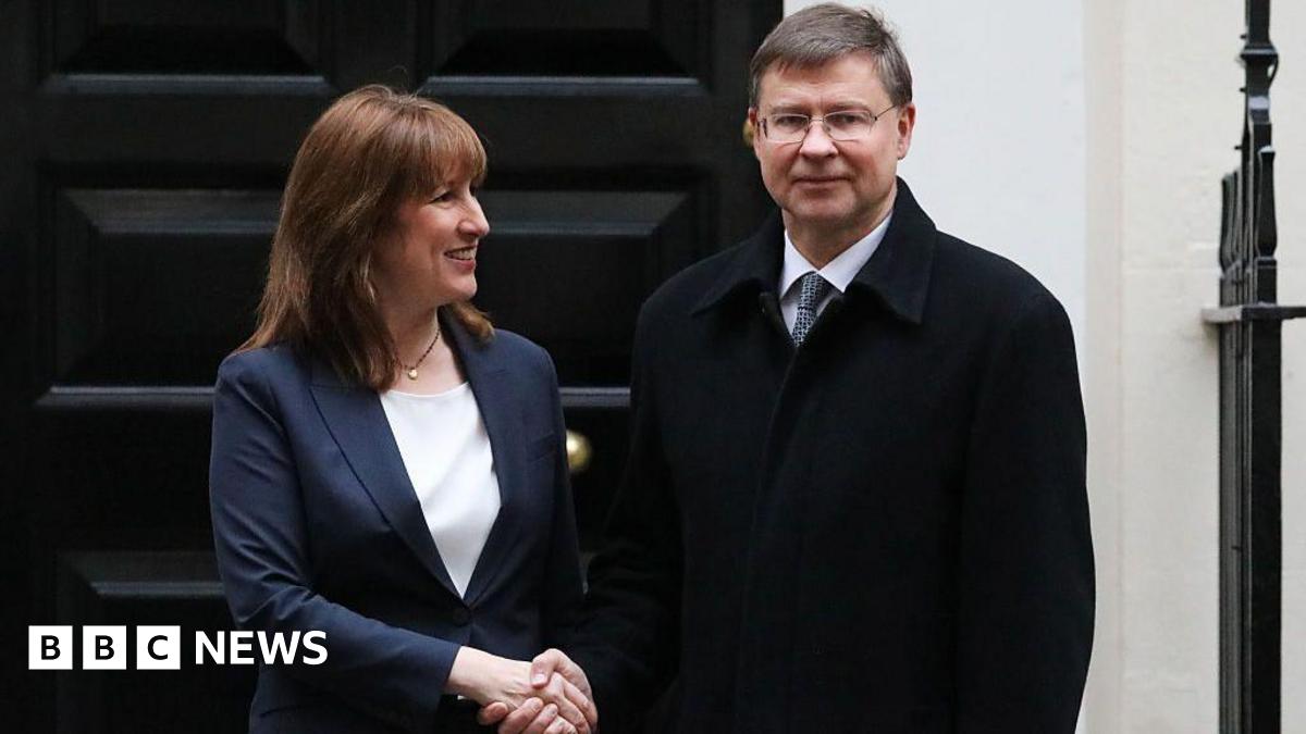Rachel Reeves, wearing a blue jacket, and Valdis Dombrovskis, wearing a black coat, shake hands while standing in front of a black door.