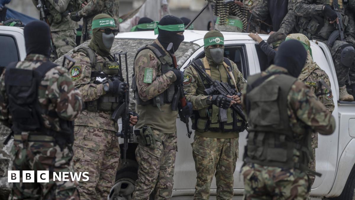 Armed members of Hamas stand in camouflage uniforms in front of a white car. Their faces are covered by balaclavas.