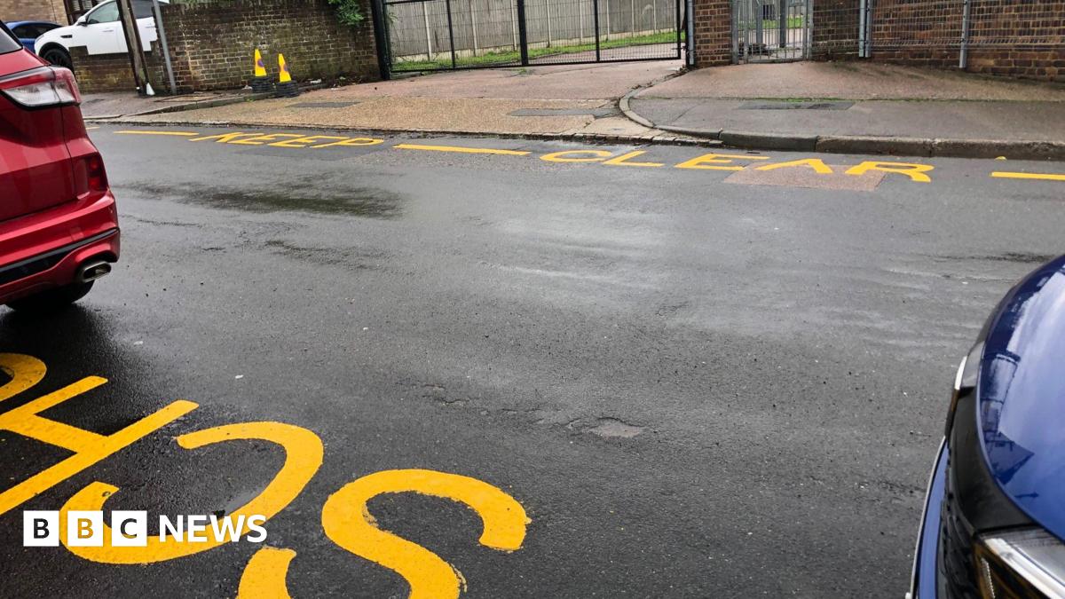 The image shows repainted yellow zigzags and 'School Keep Clear' warnings outside the former site of Halfway Houses Primary School in Sheerness, Kent. A small section of a red car can be seen in the image, and a small section of a separate blue car can...