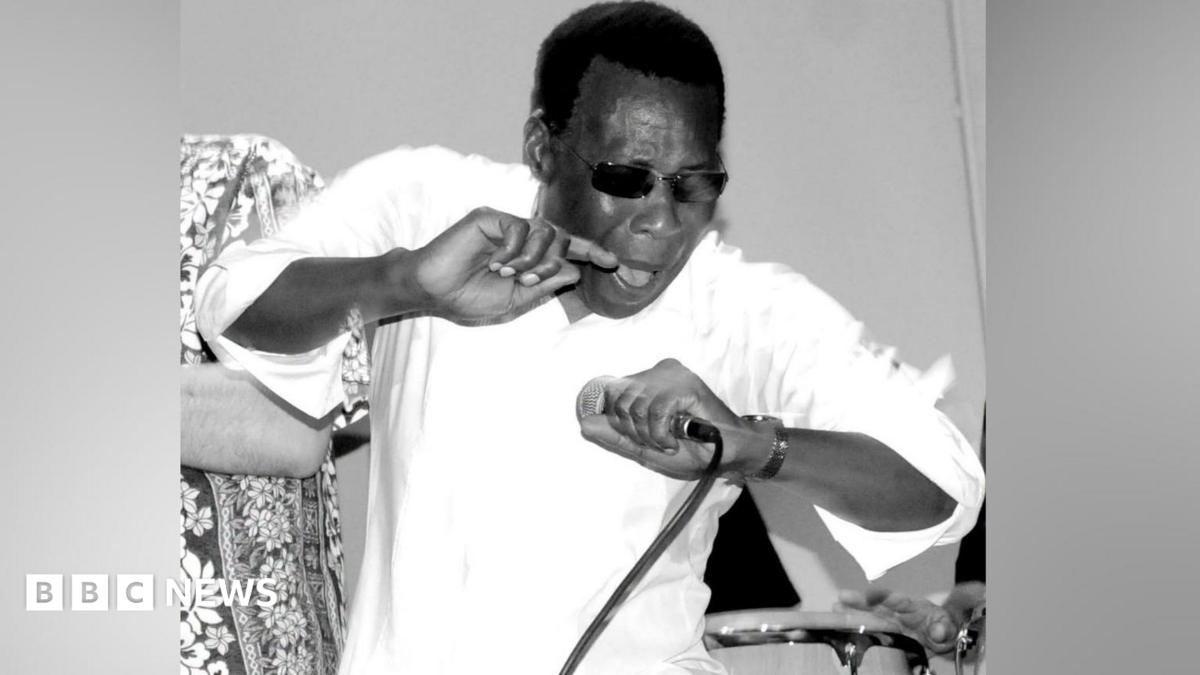Ray King in stage in an black-and-white image. He is wearing a white shirt and had dark glasses on. He is holding a microphone in his left hand. (Ảnh: BBC News)