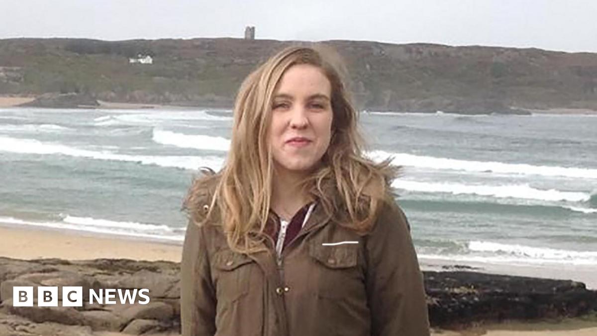 Natalie McNally. She has long blonde hair and is wearing a long khaki coat and standing on a beach.