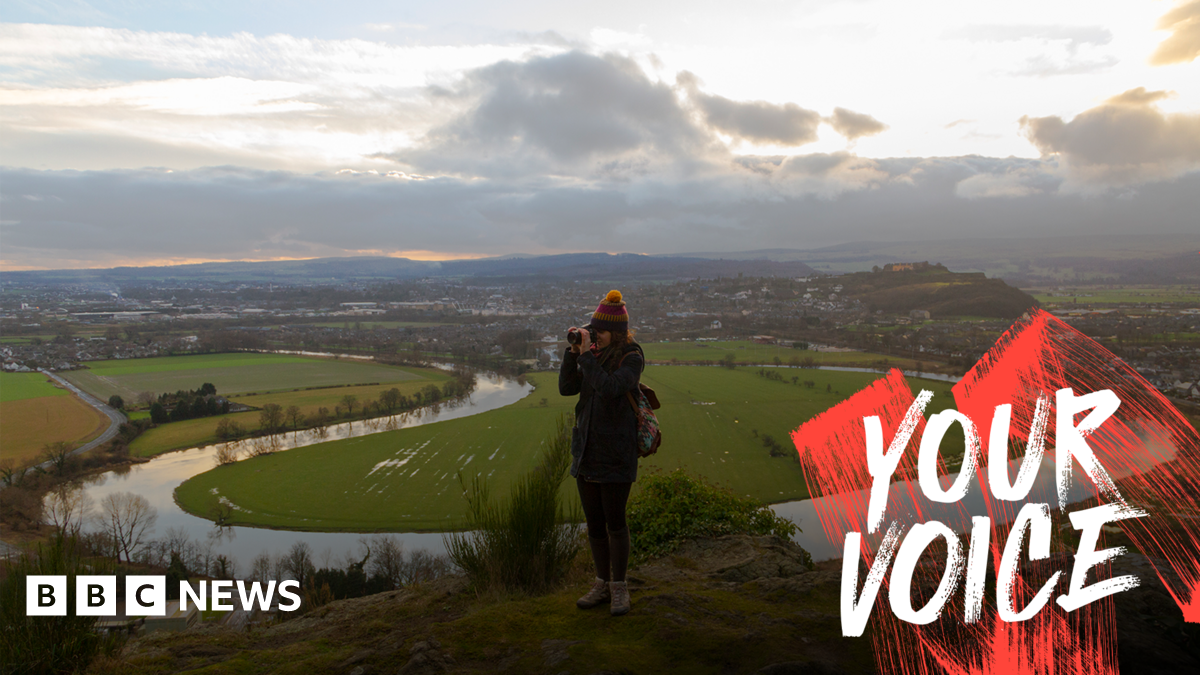 A woman is standing on top of her grassy hill. Behind her is the large meander of a river and the Scottish countryside. The sun is trying to break through the clouds. 