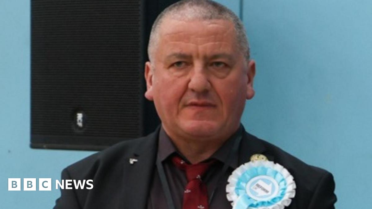 Andrew Harrison is wearing a black suit with a dark red tie with a light blue Reform rosette. He has close cropped grey hair.