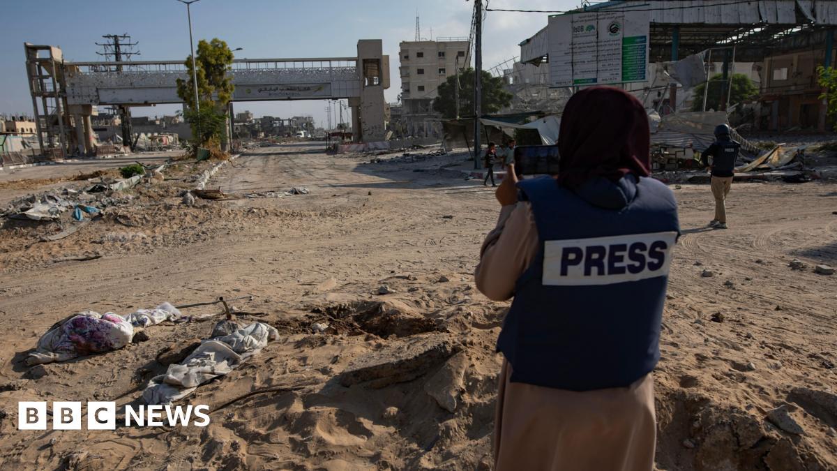 A journalist wearing a protective vest with the word "PRESS" across the back stands on a road strewn with debris in Gaza