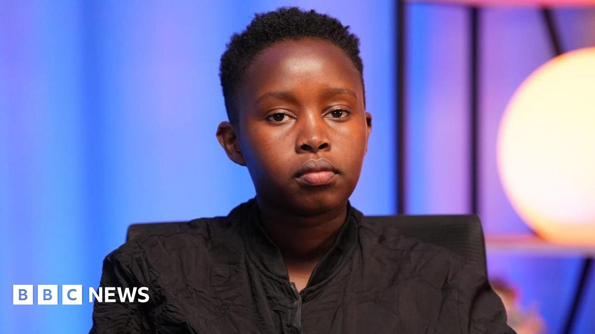 Esther Njoki stares at the camera. She's drressed in a black jacket in a purple room, with a bright, circular cream-coloured light in the background