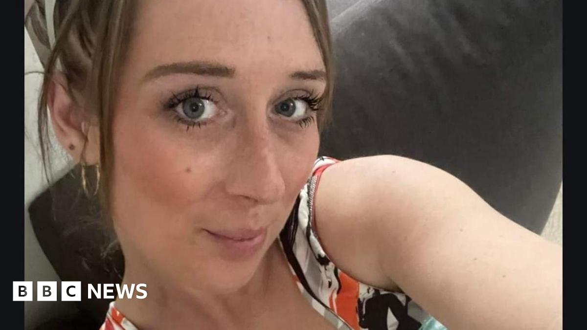 Alana Odysseos is taking a selfie wearing gold hoop earrings and an orange, white, grey and black patterned sleeveless top. She is sitting on a grey sofa.