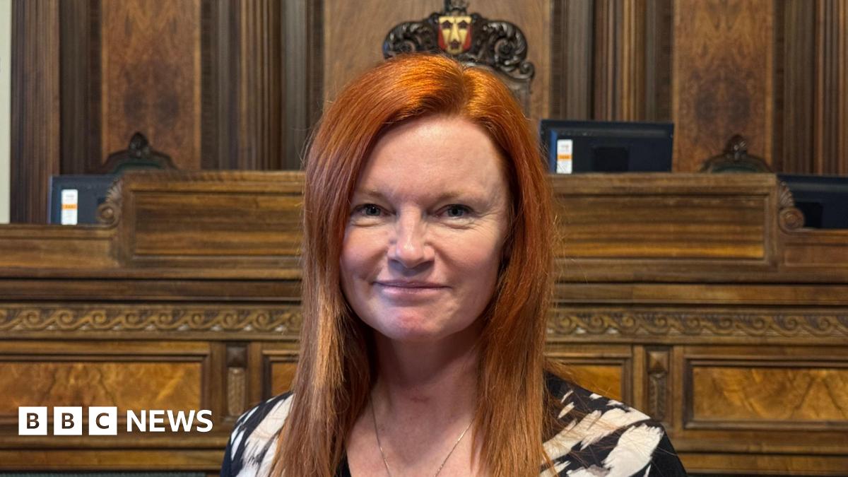 An image of a red-headed Ella Worthington, she has long hair and is smiling.  She is standing in the council chamber, with wood panelling and seating behind her.  She is wearing a black and white top and a necklace.