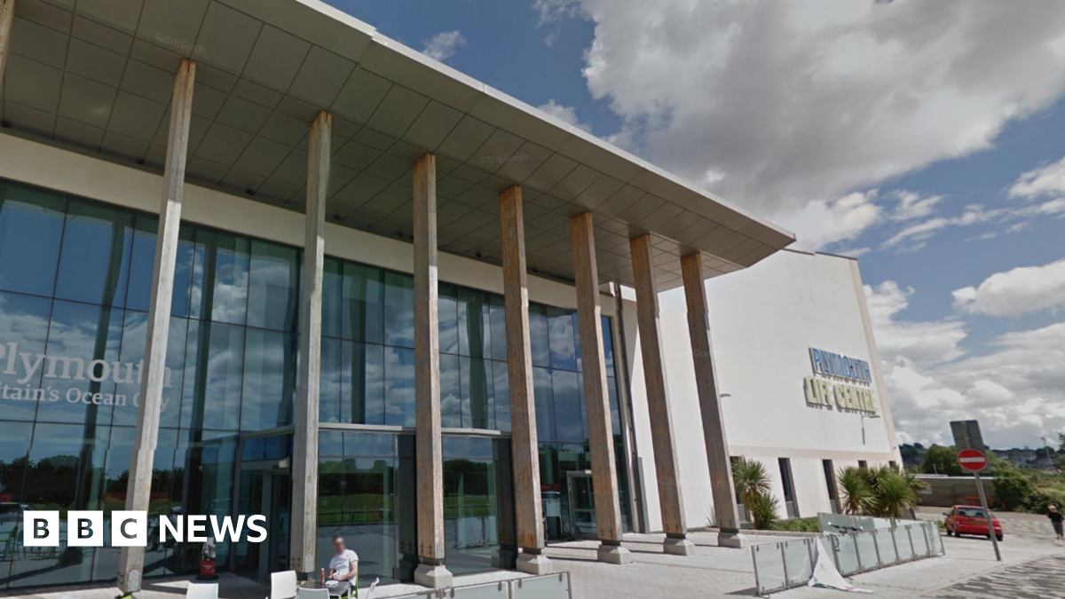 The outside of Plymouth Life Centre. It is a large glass fronted building with seven pillars propping up an extended solid awning. There is cloudy sky in the background of the image.