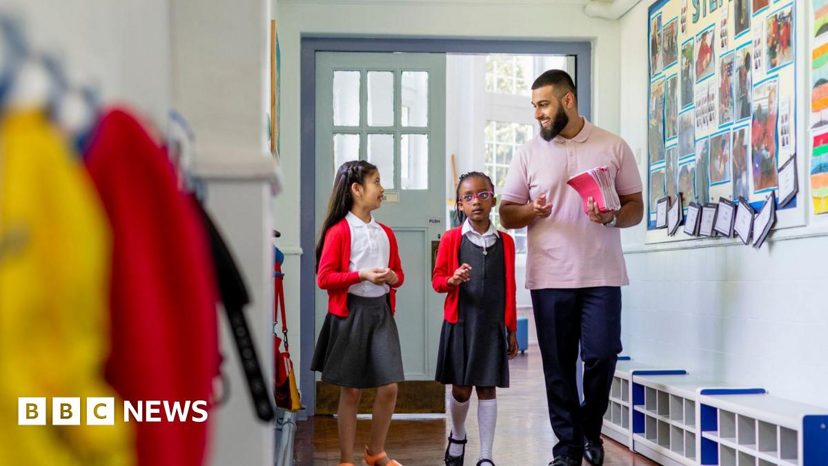 A stock image of two female primary school pupils, both in red cardigans, walking along a school corridor with their male teacher who is wearing a pink polo shirt and carrying a stack of pink notebooks.