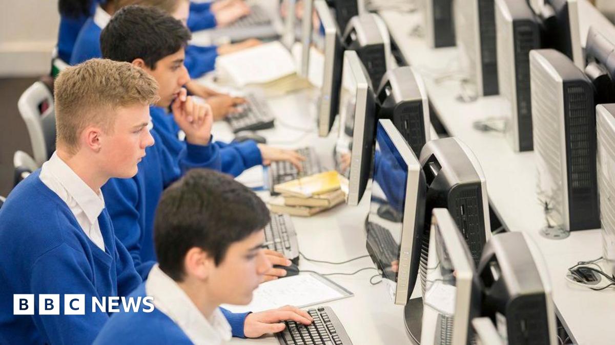 Three boys wearing blue school jumpers and white shirts sit in front of computers in a classroom. They are looking straight ahead at their screens