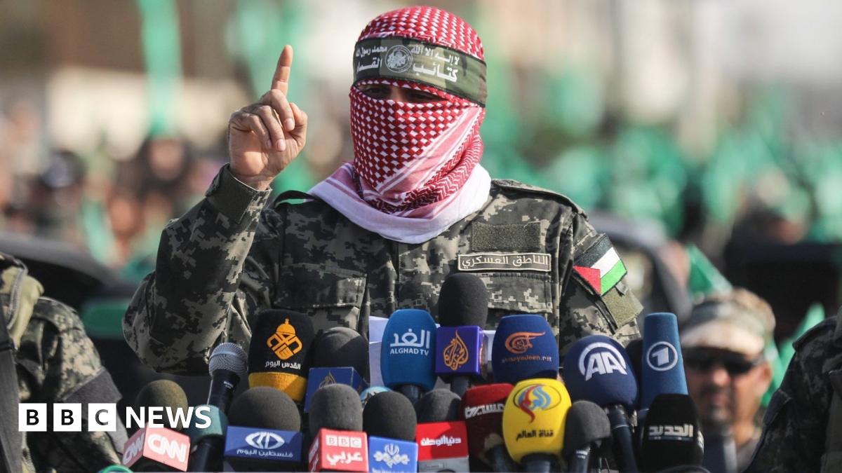 Abu Obeida speaks behind a number of microphones with logos of world media while wearing a scarf over his face, flanked by two Hamas fighters.