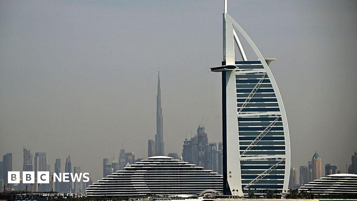 Buildings on the Dubai skyline, with tall towers seen in the background and the sail-shaped hotel the Burj Al Arab in the foreground, against a grey sky 