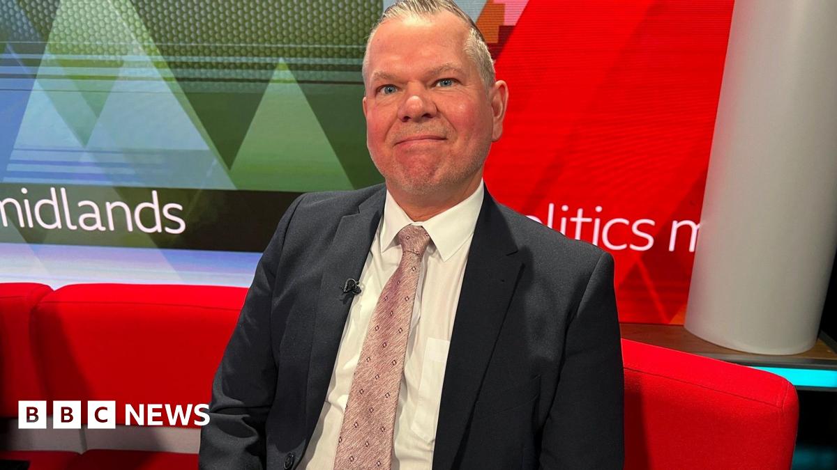A man wearing a suit and pink tie sits on red sofa looking at the camera. A screen can be seen behind him.
