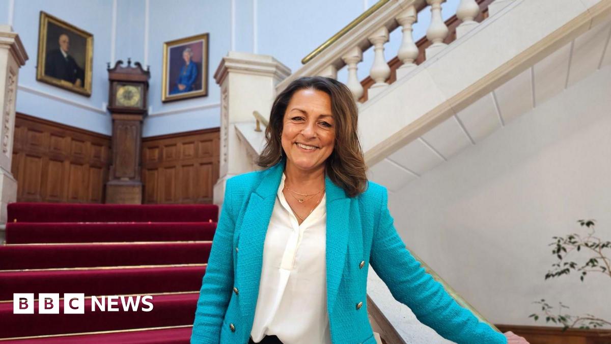Linden Kemkaran stands on a staircase and holds the bannister with one hand. She is wearing a blue suit jacket and smiling at the camera. Behind her the staircase is covered in a red carpet and a grandfather clock stands on a landing area.