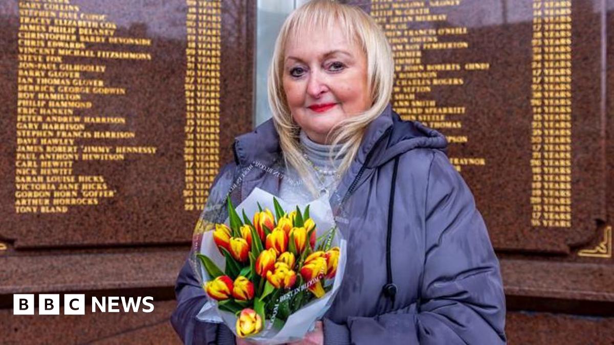 Jenni Hicks, with blonde hair, red lipstick and wearing a blue coat, stands before the Hillsborough memorial which has the names of the 97 engraved in golden lettering, holding a bunch of tulips. 