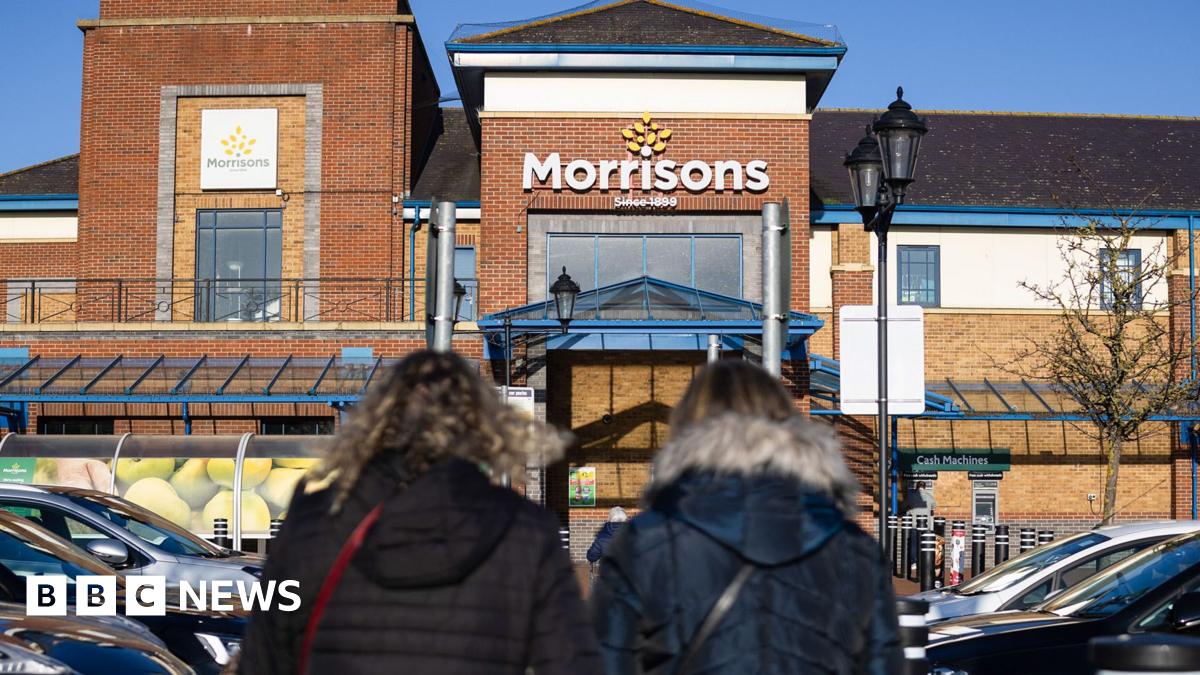 Two women in coats walking into a large Morrisons store with the logo above the entrance