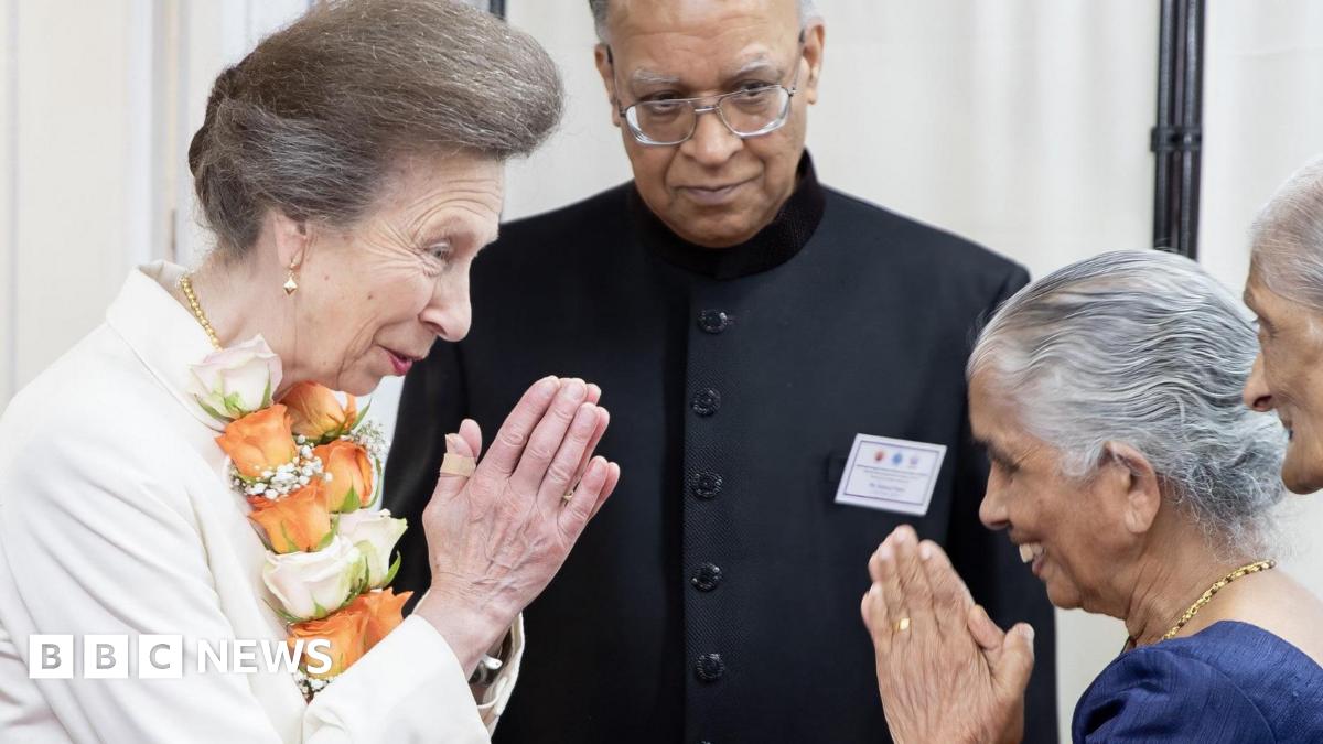 A woman in a white top in an orange garland puts her hands together in prayer next to a man in a black suit and two women in Hindu dress. 