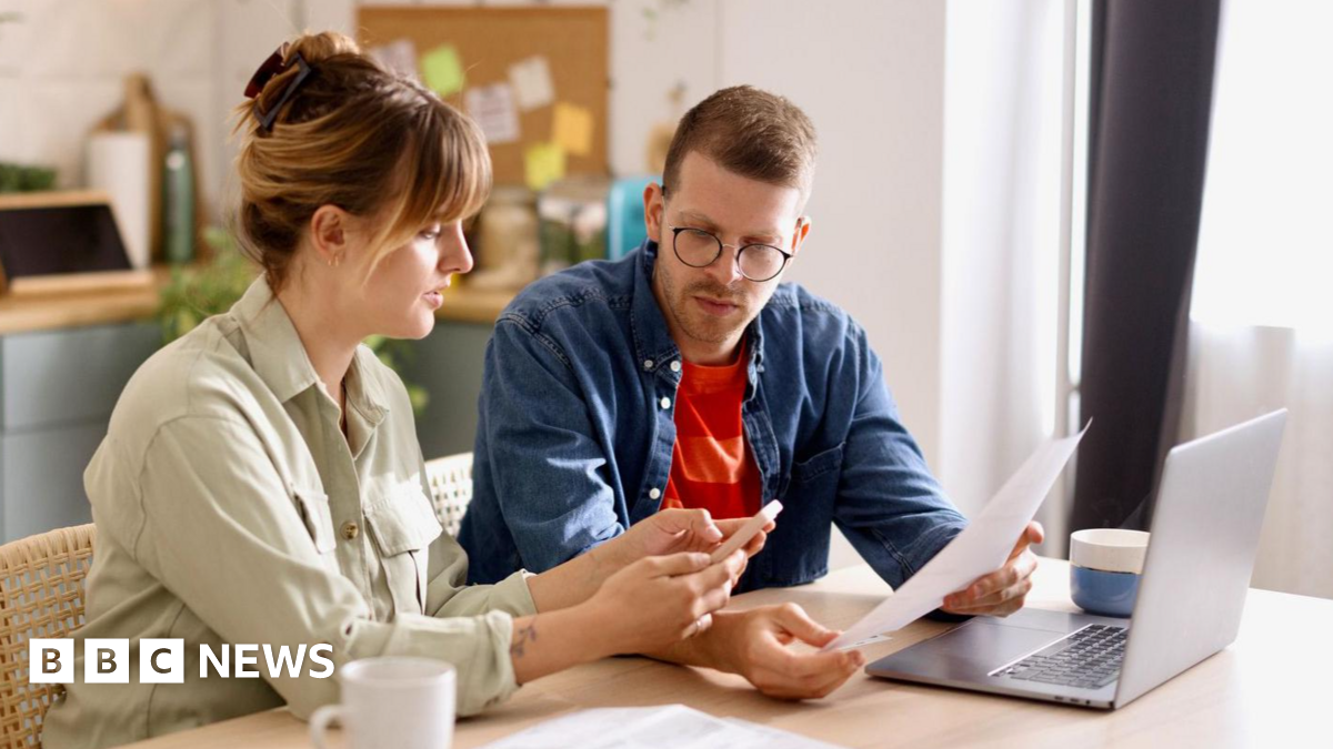 Couple go through finances on the kitchen table with bills, a laptop and a mug in front of them.