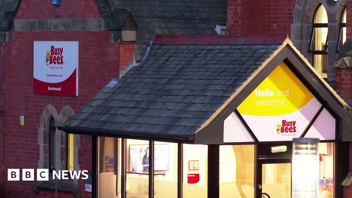 A large red brick building with a sloped roof and arched windows. We can see a 'Hello and welcome to Busy Bees' sign. The lights are on bright. It is evening time so the rest of the building is slightly dark.  