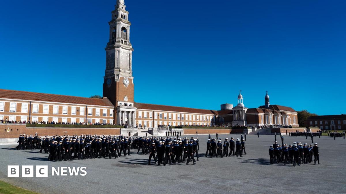A view of the front of Royal Hospital School. There is a long red building with a large spire in the middle of it. Students wearing uniforms similar to those worn by the navy walk in groups in a large quad in front of the school building.