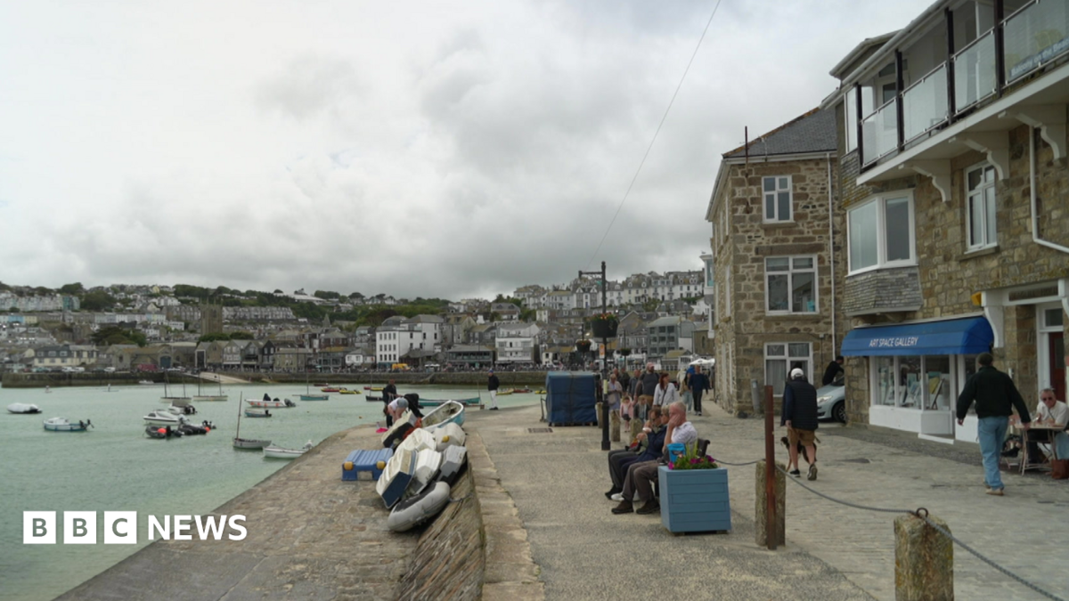 A harbour scene shows a stone promenade lined with buildings, with small boats floating on calm water and a town spread across the hillside in the background under a cloudy sky. People are sitting and walking along the waterfront near stacked dinghies,...