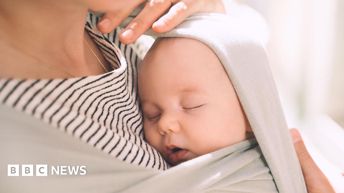 An unidentifiable mum, wearing a stripey top, holds her young baby wrapped tightly in a light grey sling close to her chest.
