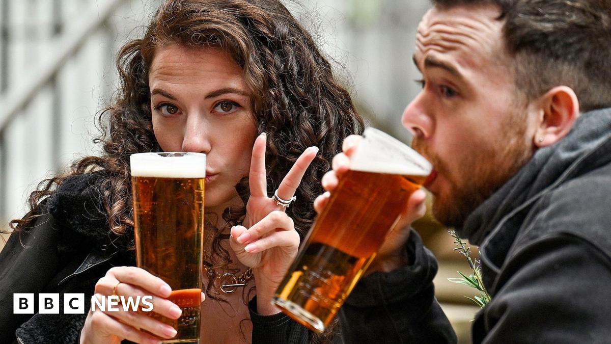 A woman and a man sitting outdoors holding pints of Lager. She is making a peace sign and he is drinking, in a casual seating area with steps and railings in the background