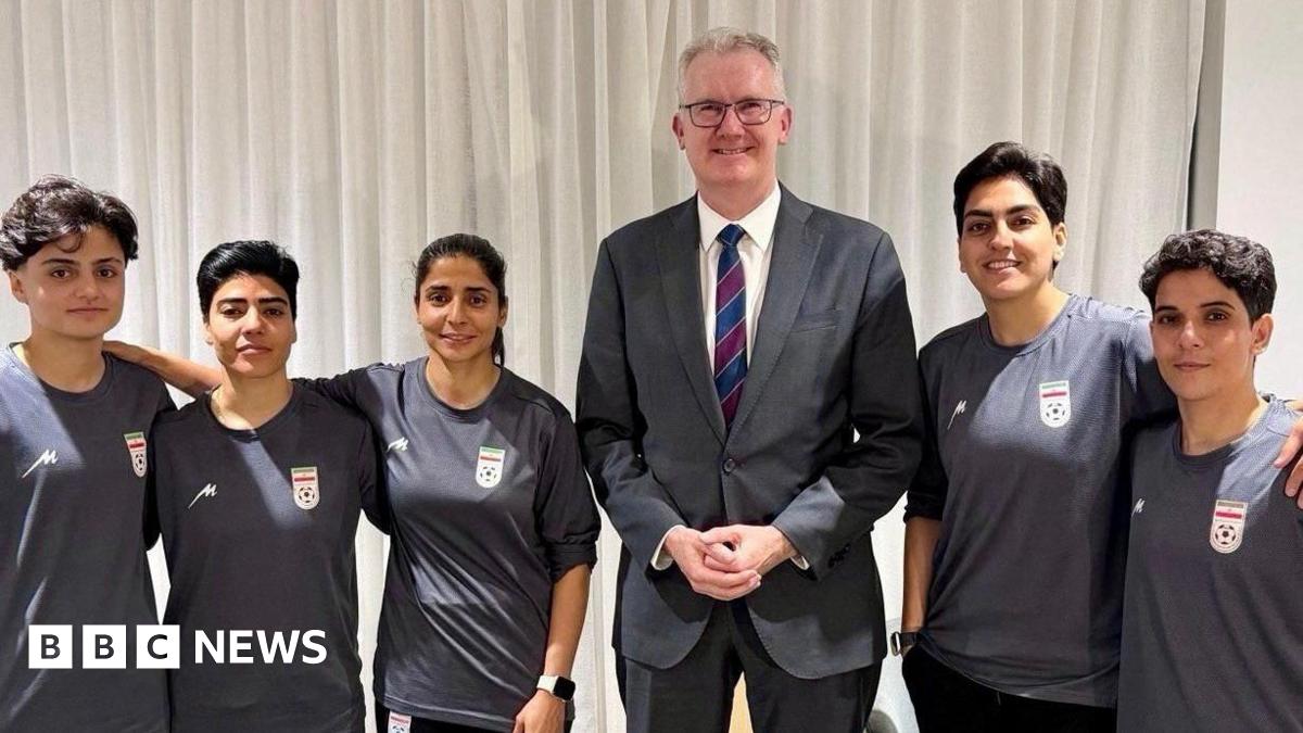 Australian Home Affairs Minister Tony Burke, wearing a grey suit, poses with the five Iranian football players who have been granted humanitarian visas. They are all smiling and wearing grey football jerseys.