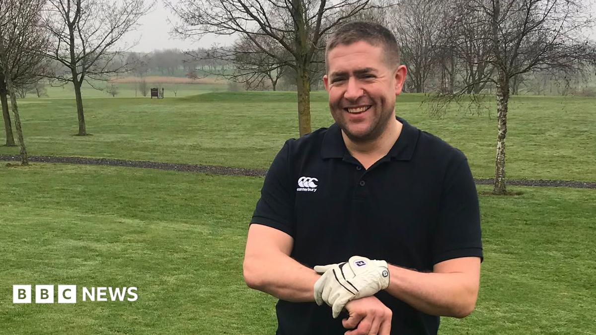 A man wearing a black polo-shirt while smiling at the camera. He is wearing white gloves while holding onto a golf club. He is standing in a field 