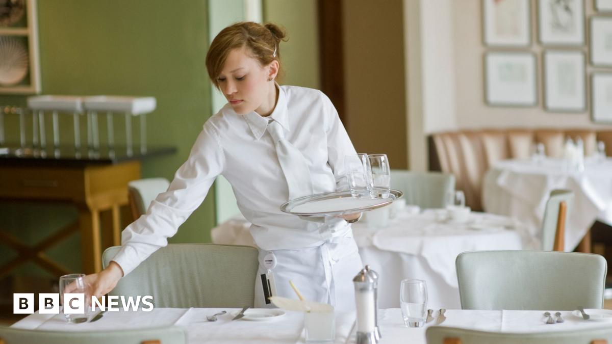 A waitress dressed in a white shirt, white tie and white apron sets out water glasses in restaurant