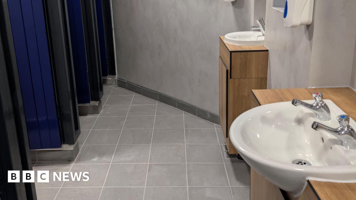 A clean, modern public restroom with a tiled grey floor and smooth light‑grey walls. Along the right side, there are two white sinks mounted on wooden cabinets, each fitted with chrome taps. A soap dispenser is mounted on the wall above the sinks. On t...