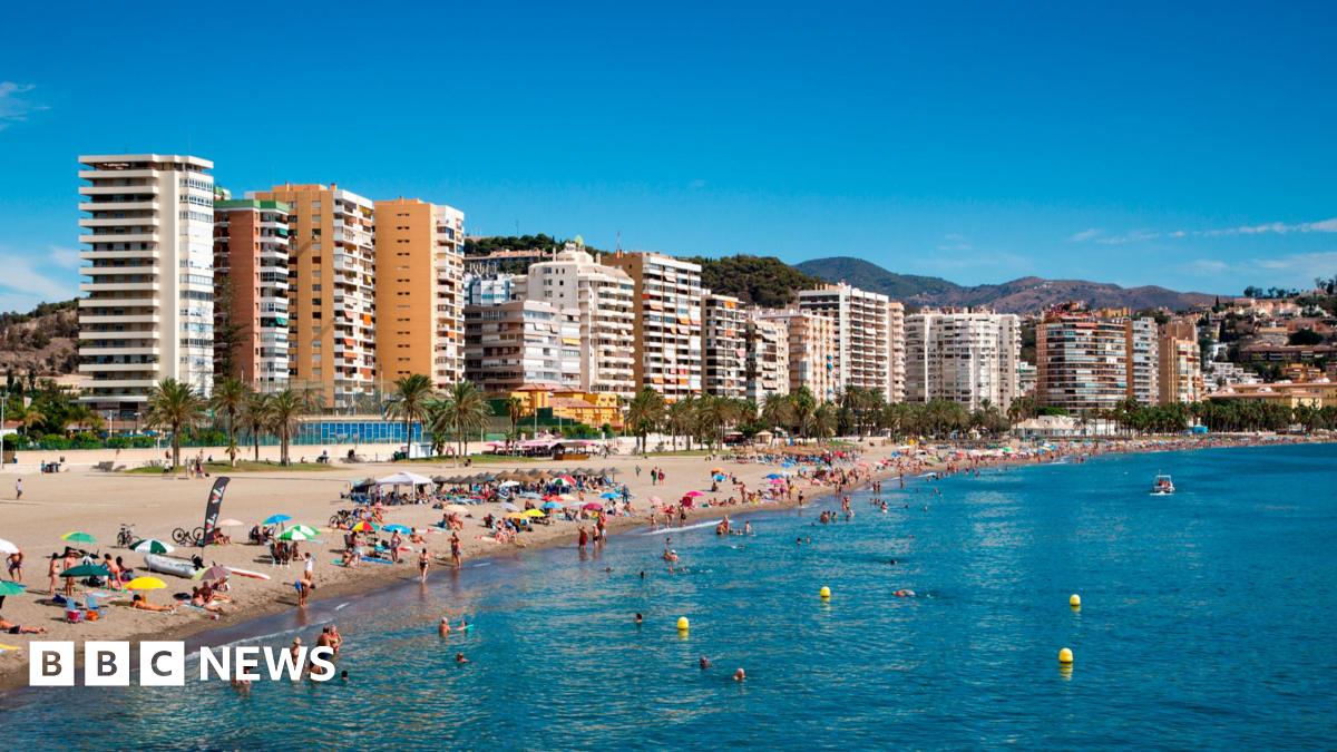 A sunny beach scene with people sunbathing on the shore and some people swimming. There are high-rise buildings in the background.