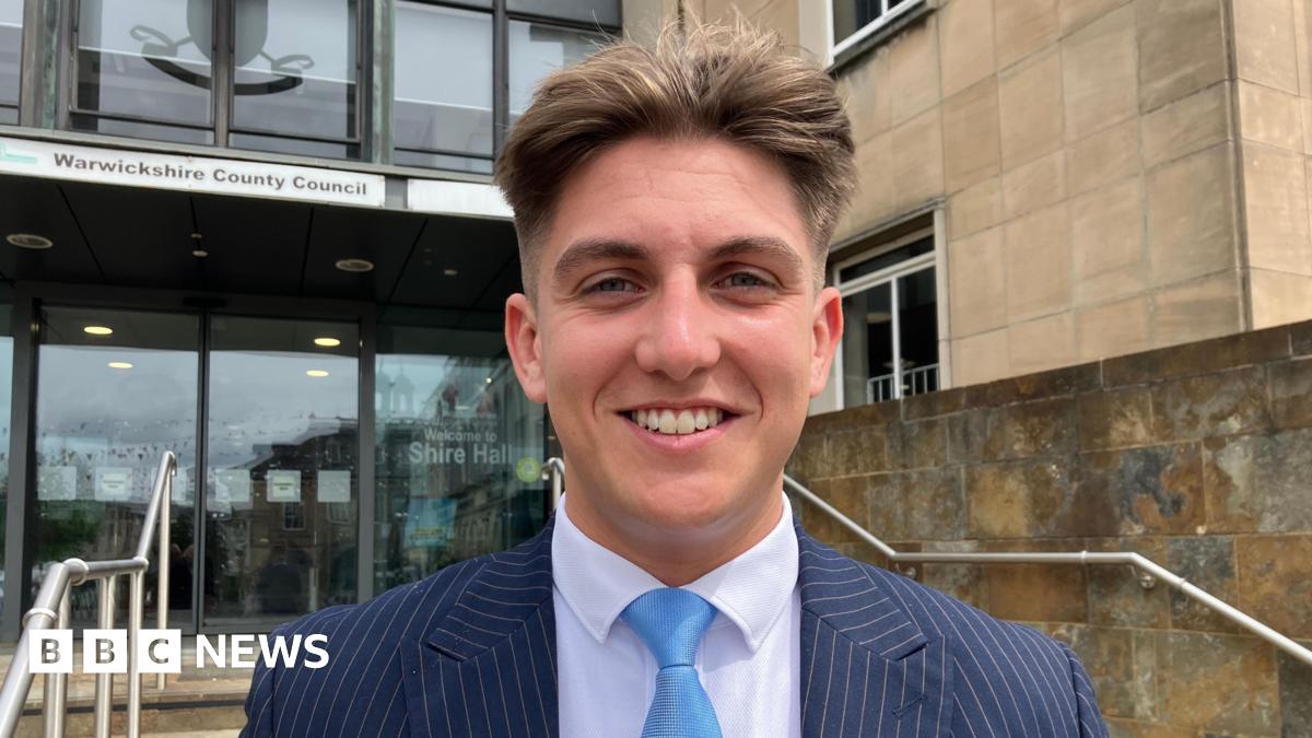 George Finch stands outside the entrance to Warwickshire County Council. He wears a striped suit and a blue tie and is smiling at the camera. He is clean shaven and has short dark hair.