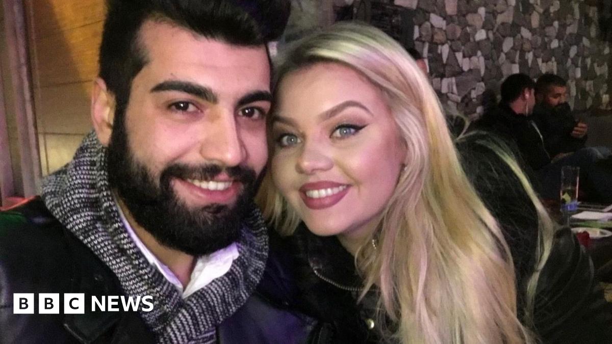A couple smile at the camera. She has blonde hair and blue eyes. He has dark hair and a beard, and brown eyes. They are taking a selfie inside a restaurant or bar.